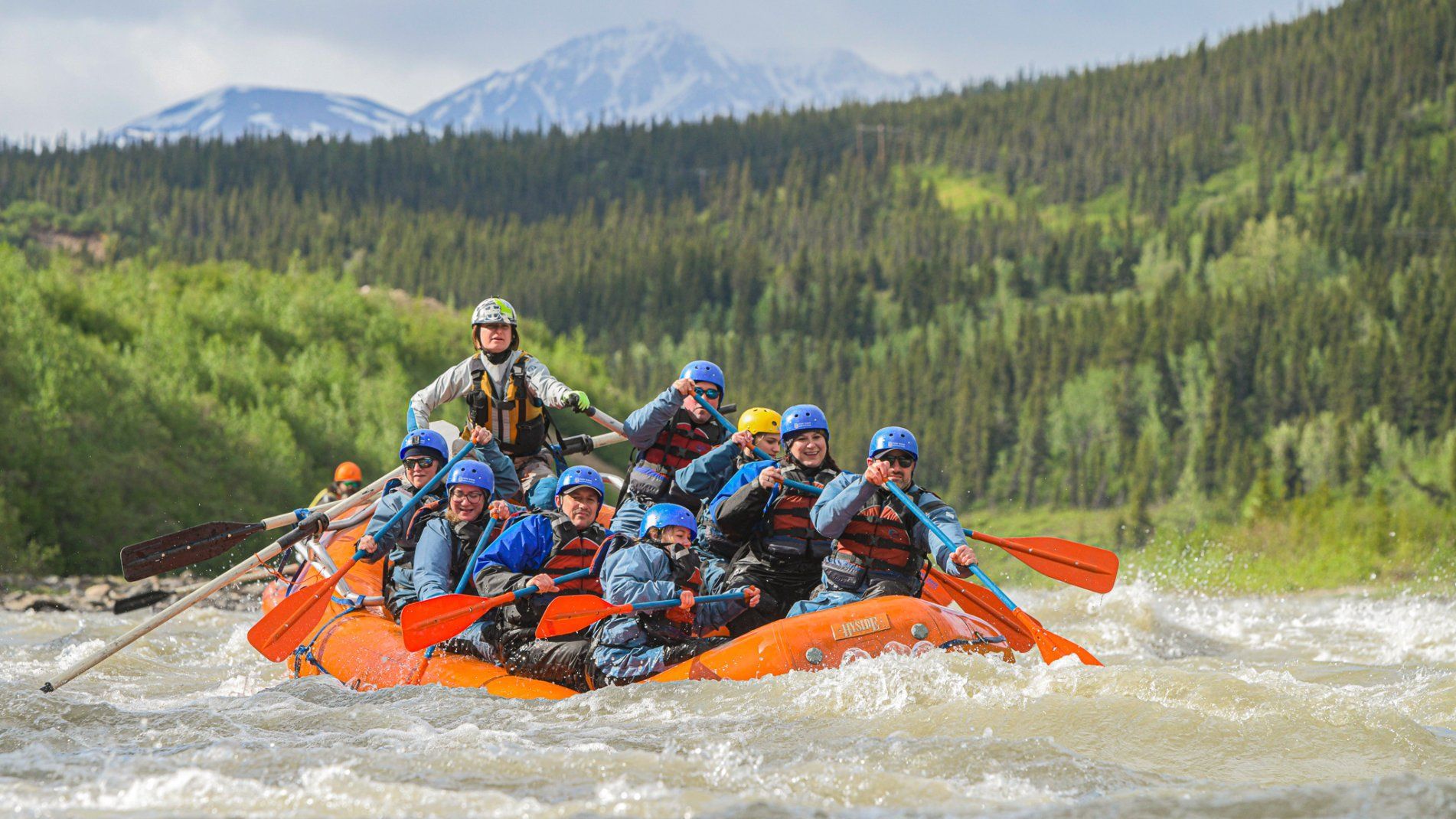 Group of rafters in orange boats paddling through whitewater with forested mountains in the background