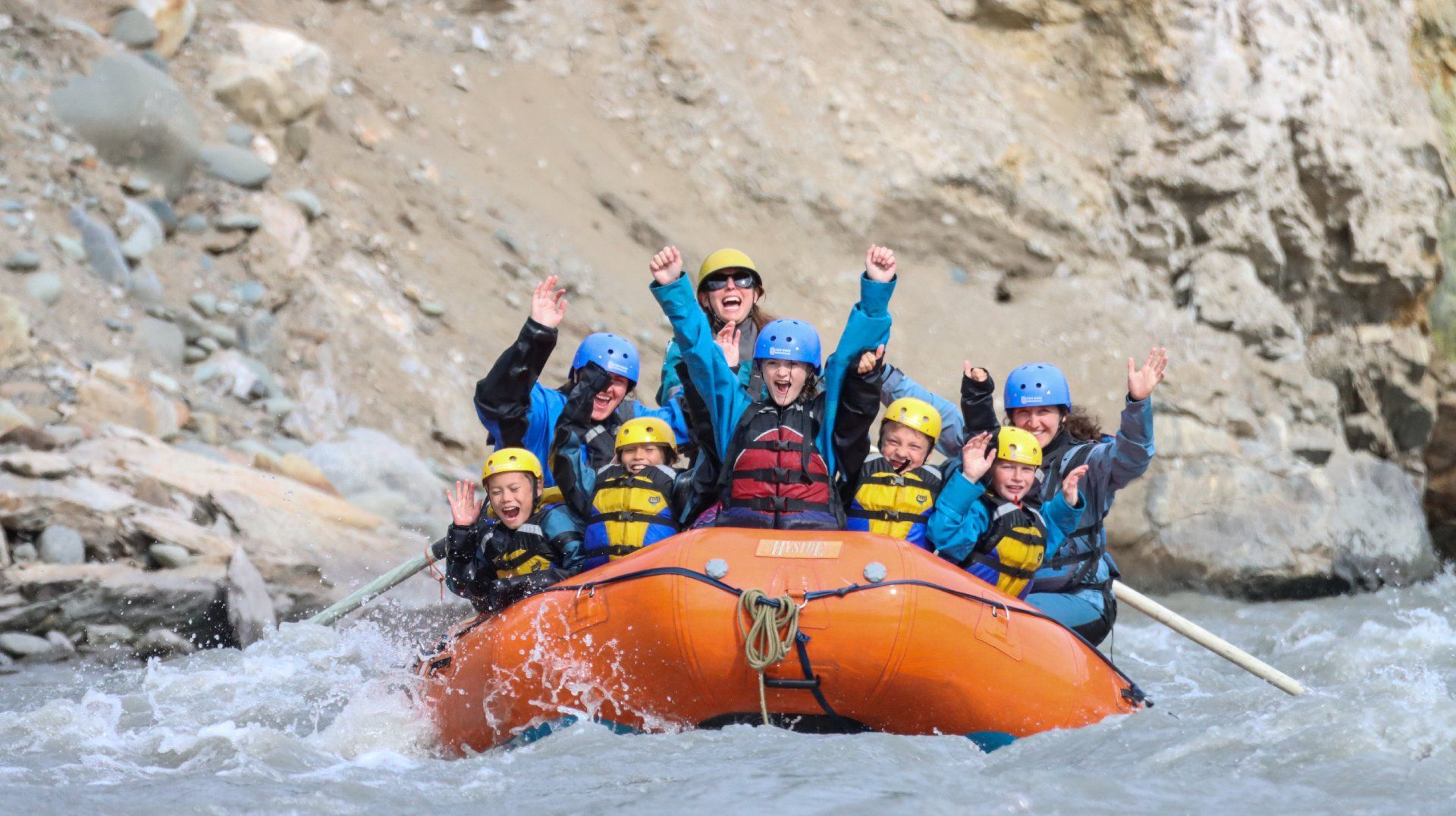 Group rafting through whitewater in an orange raft, paddles raised, between rocky canyon walls