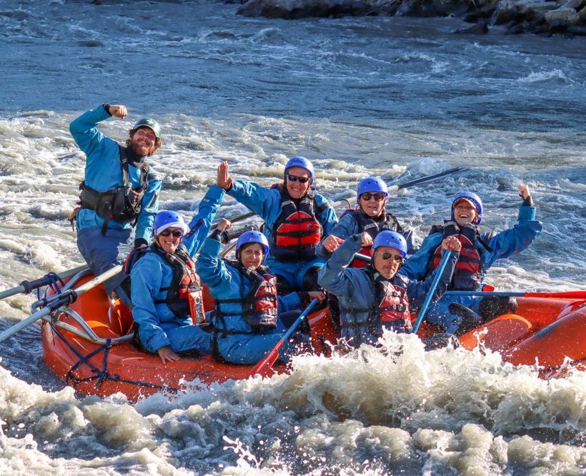 People whitewater rafting in a red inflatable boat, splashing through rough river rapids with blue helmets and life vests
