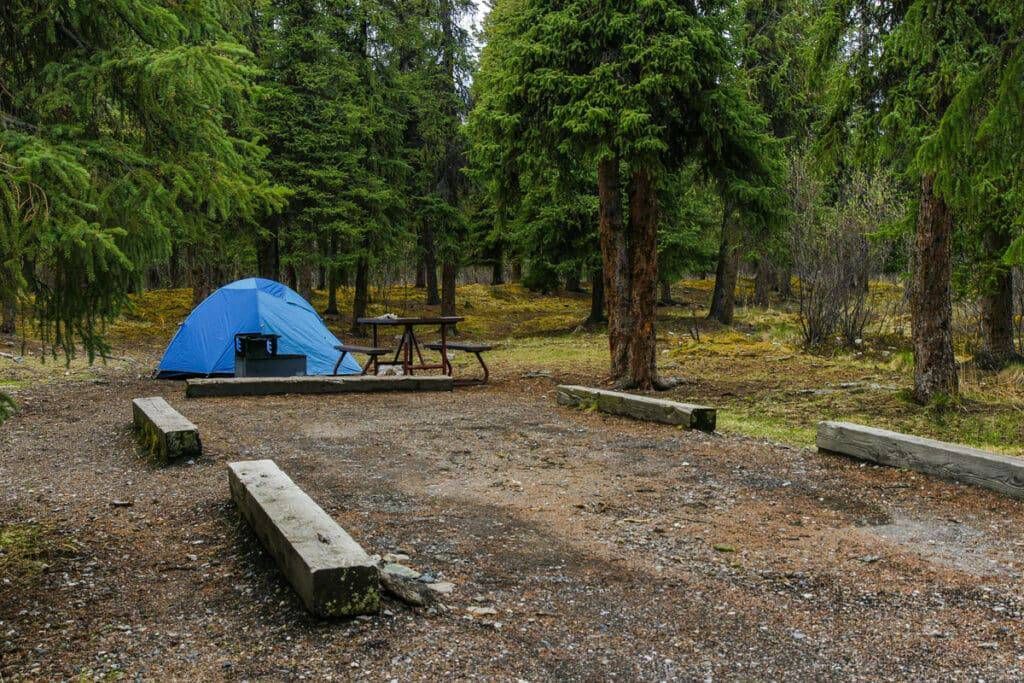 Rainy campsite with a blue tent, picnic table, and benches among trees