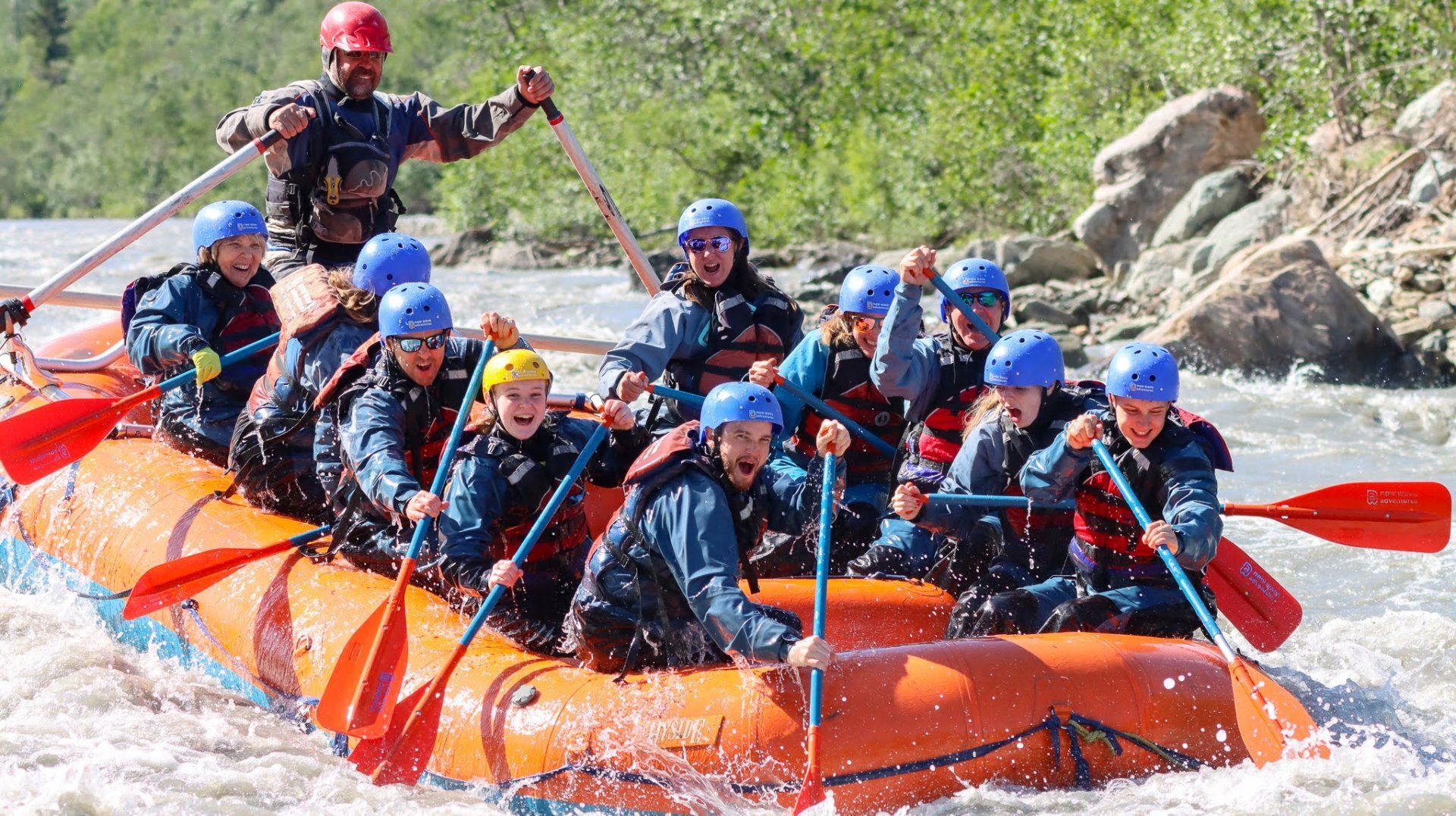 Group whitewater rafting in an orange inflatable raft through rough rapids, with people in blue helmets paddling.