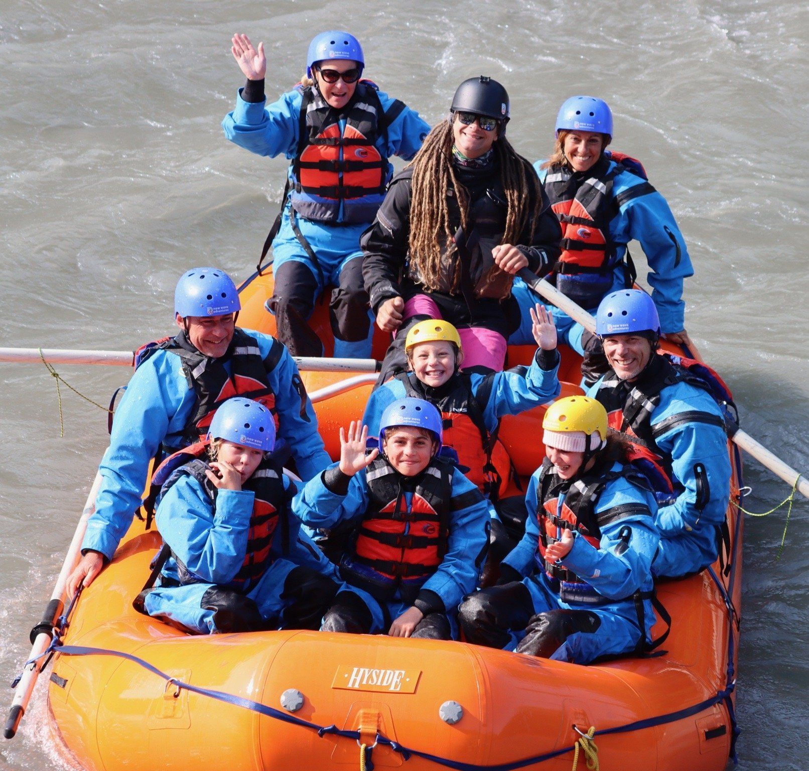 People in blue helmets and red life jackets rafting in an orange raft on rough water, one waving.