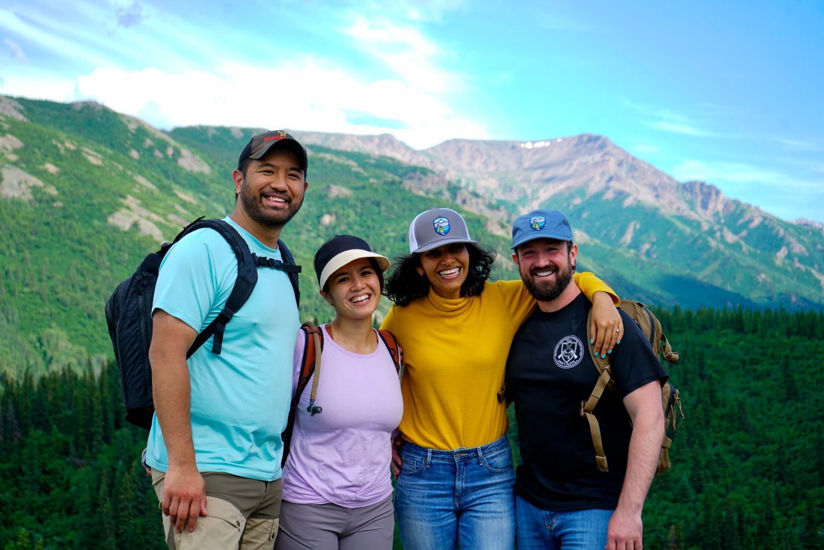 Four hikers smiling on a mountain trail with green hills and rocky peaks in the background