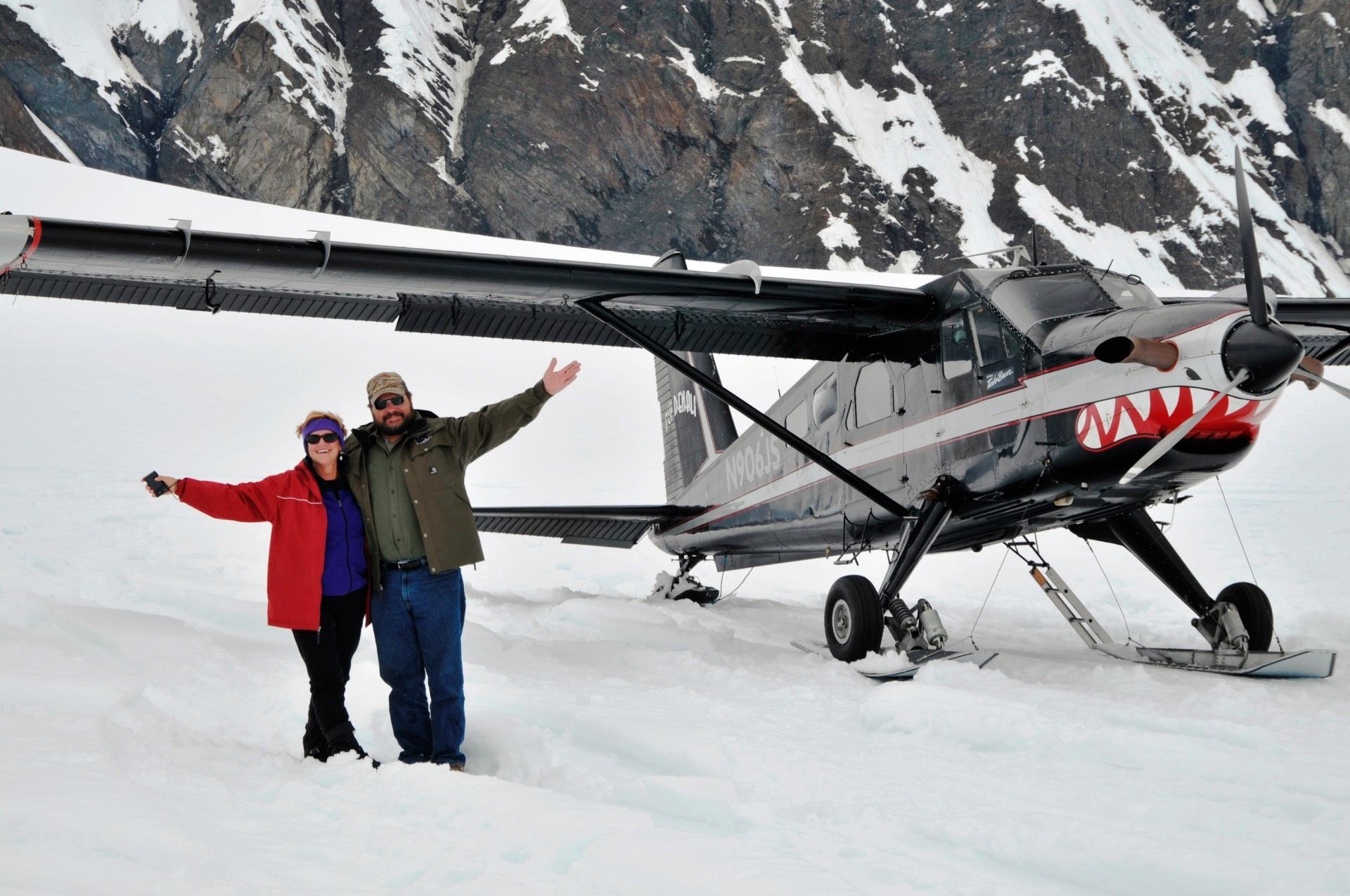 Two people posing beside a small prop plane on snow-covered mountains, one in red and one in green.
