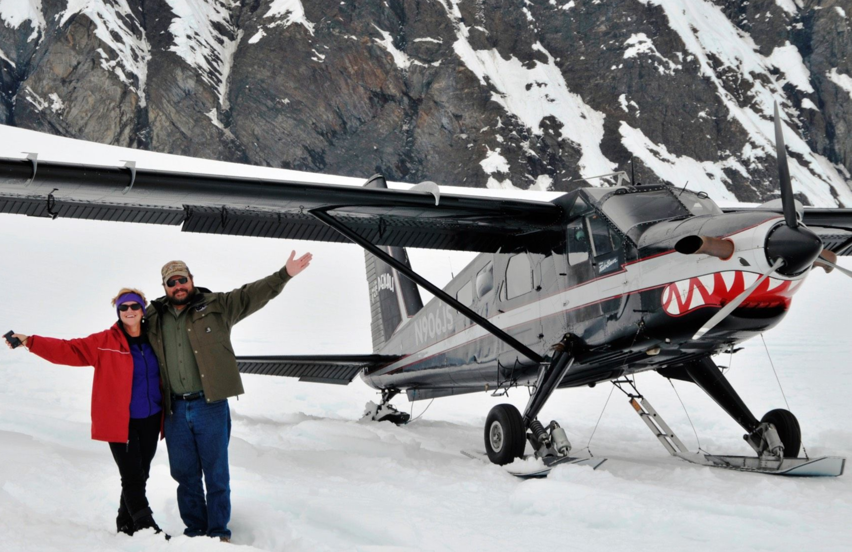 Two people posing beside a small bush plane on snowy ground with mountains behind them