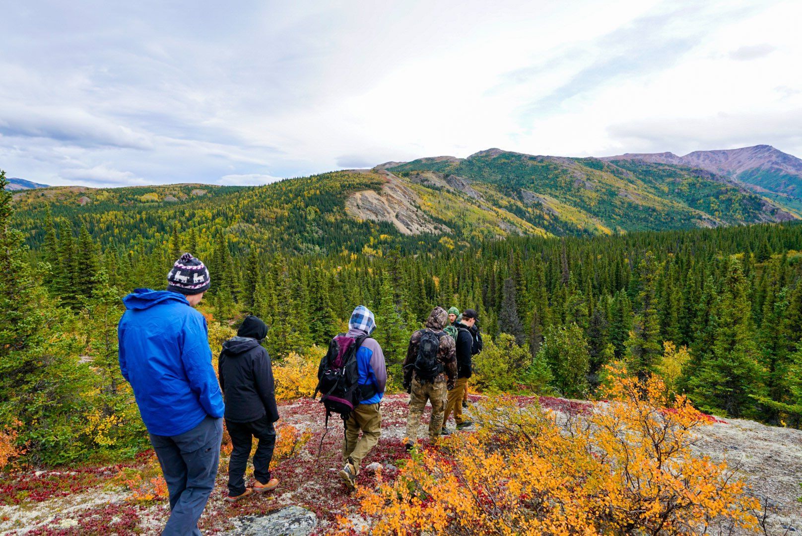 Hikers in jackets walking a mountain trail with autumn foliage and forested peaks in the background