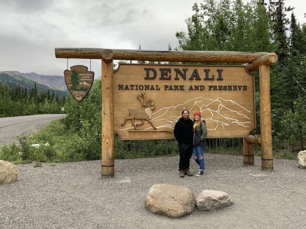 Two people standing at the Denali National Park and Preserve entrance sign with mountains in the background