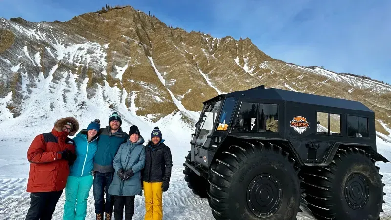 Five people in winter gear stand beside a large black off-road vehicle on a snowy mountain slope.