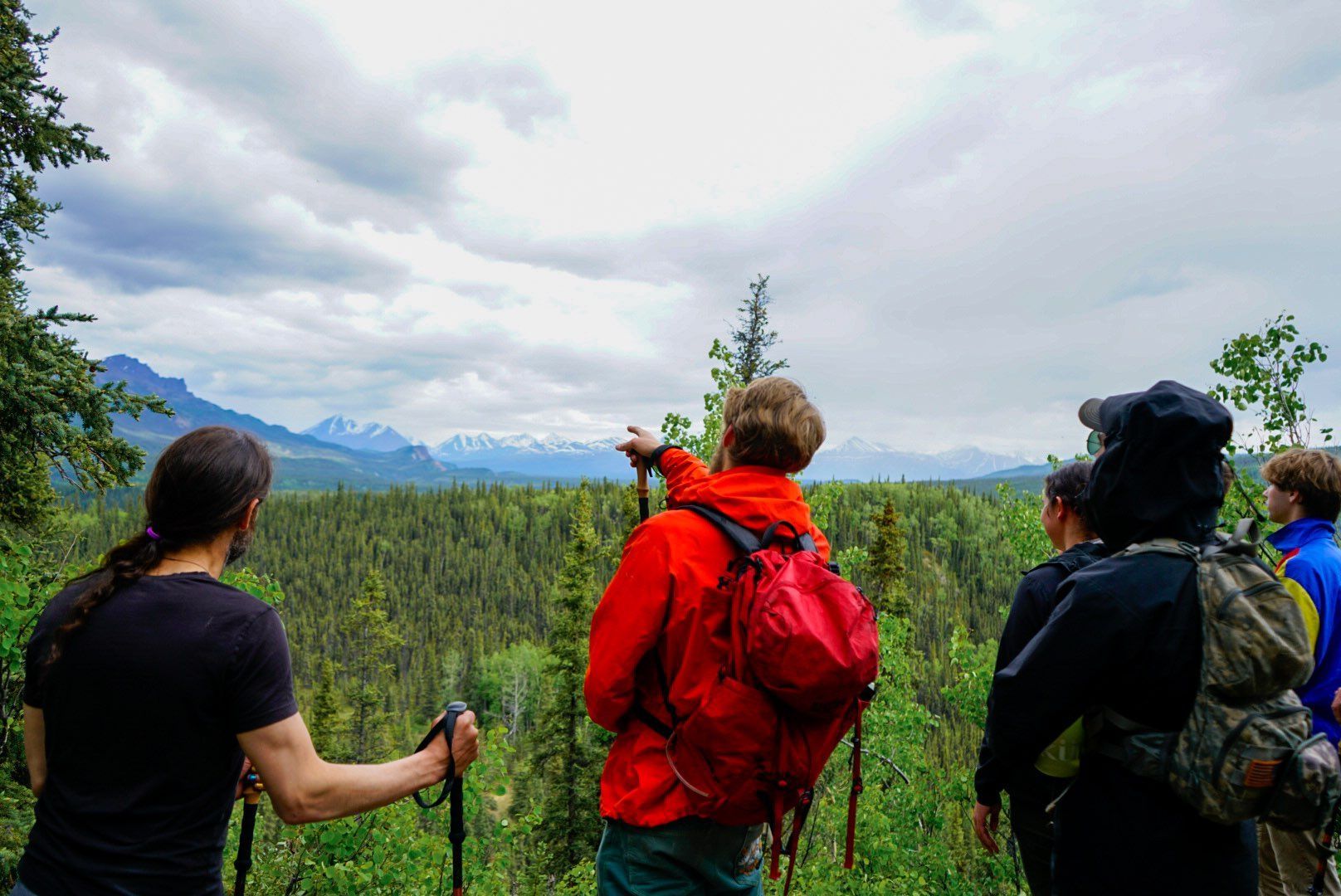 Three hikers with backpacks overlooking a green valley under cloudy skies