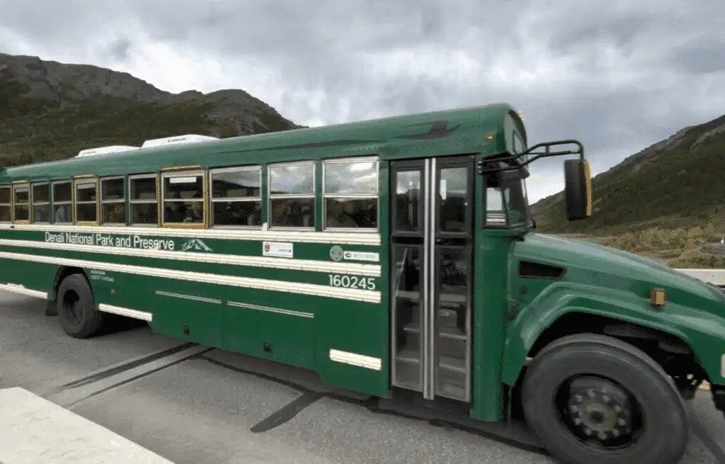 Green school bus on a mountain road, viewed from the side against cloudy hills.