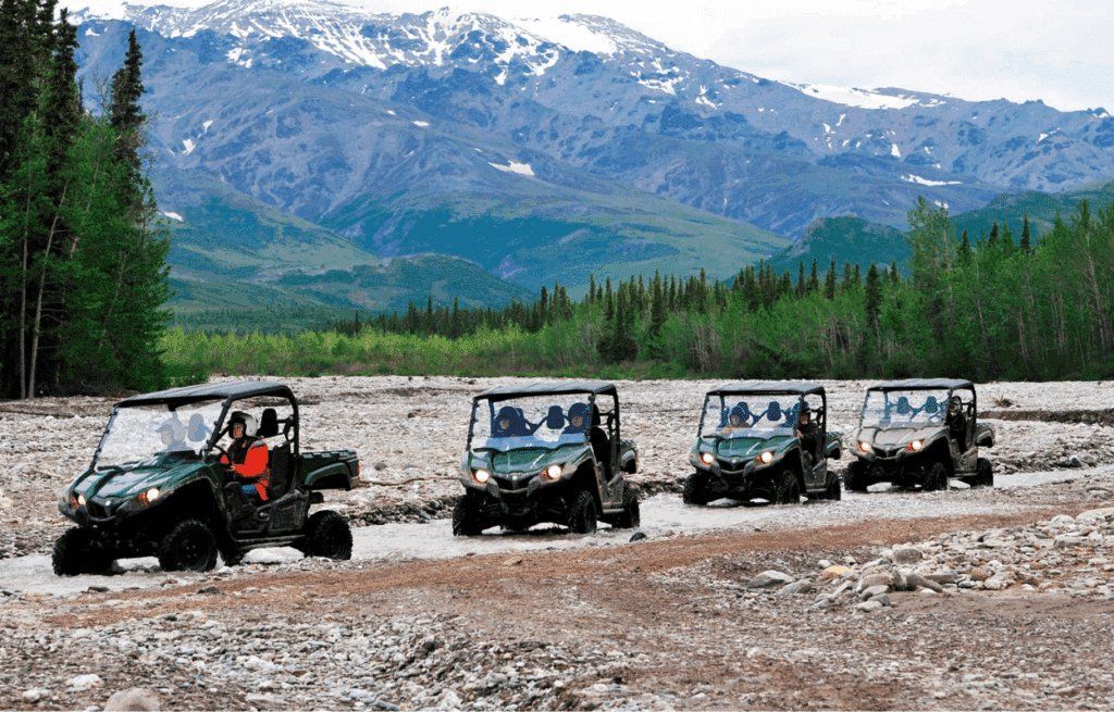 Four off-road vehicles drive through a rocky riverbed with snowy mountains in the background.