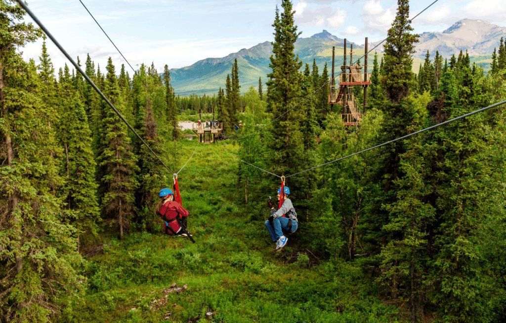Two people zipline through a green forest with mountains in the background.