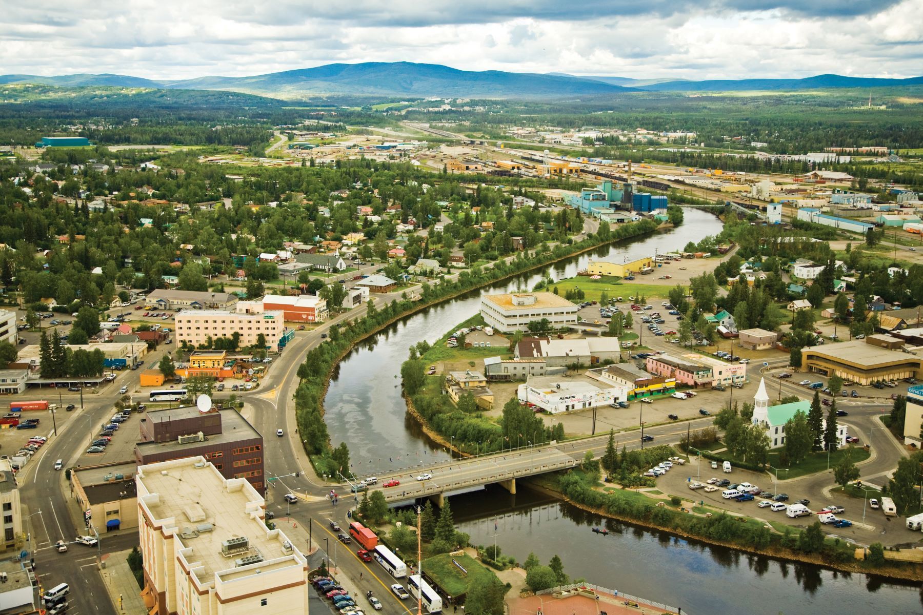 Aerial view of a city with a winding river, bridge, roads, buildings, and distant hills under cloudy skies