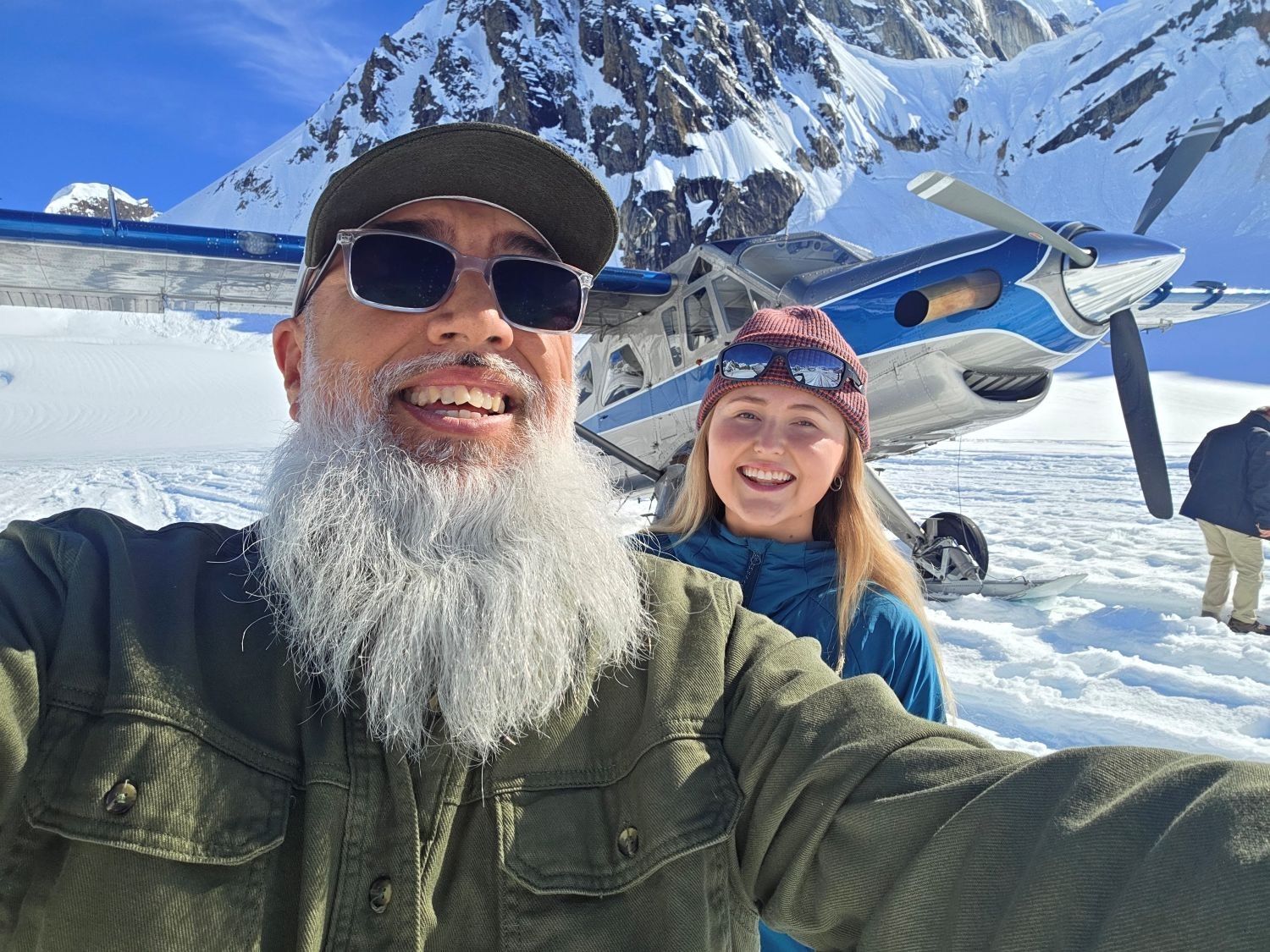 Two smiling people taking a selfie in snowy mountains with a small airplane behind them