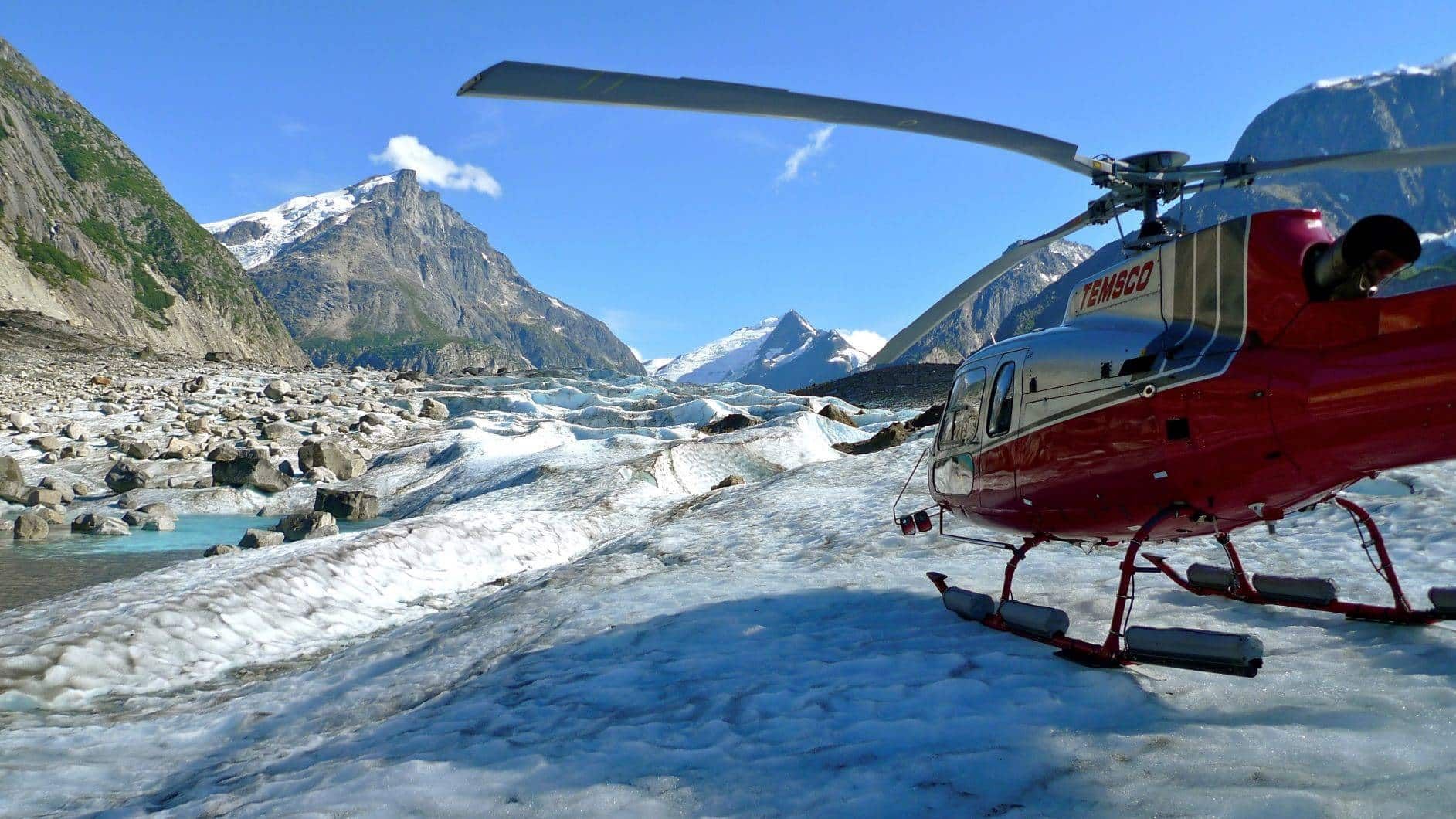 Red helicopter on snowy mountain landing area beside a rushing river and rocky peaks under a blue sky