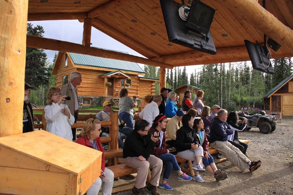 People seated under a wooden shelter watching an outdoor event near log cabins and ATVs