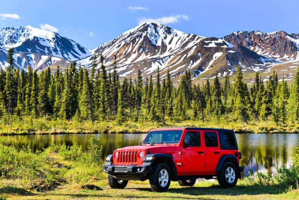 Red Jeep parked by a mountain lake with snowcapped peaks and pine trees in the background