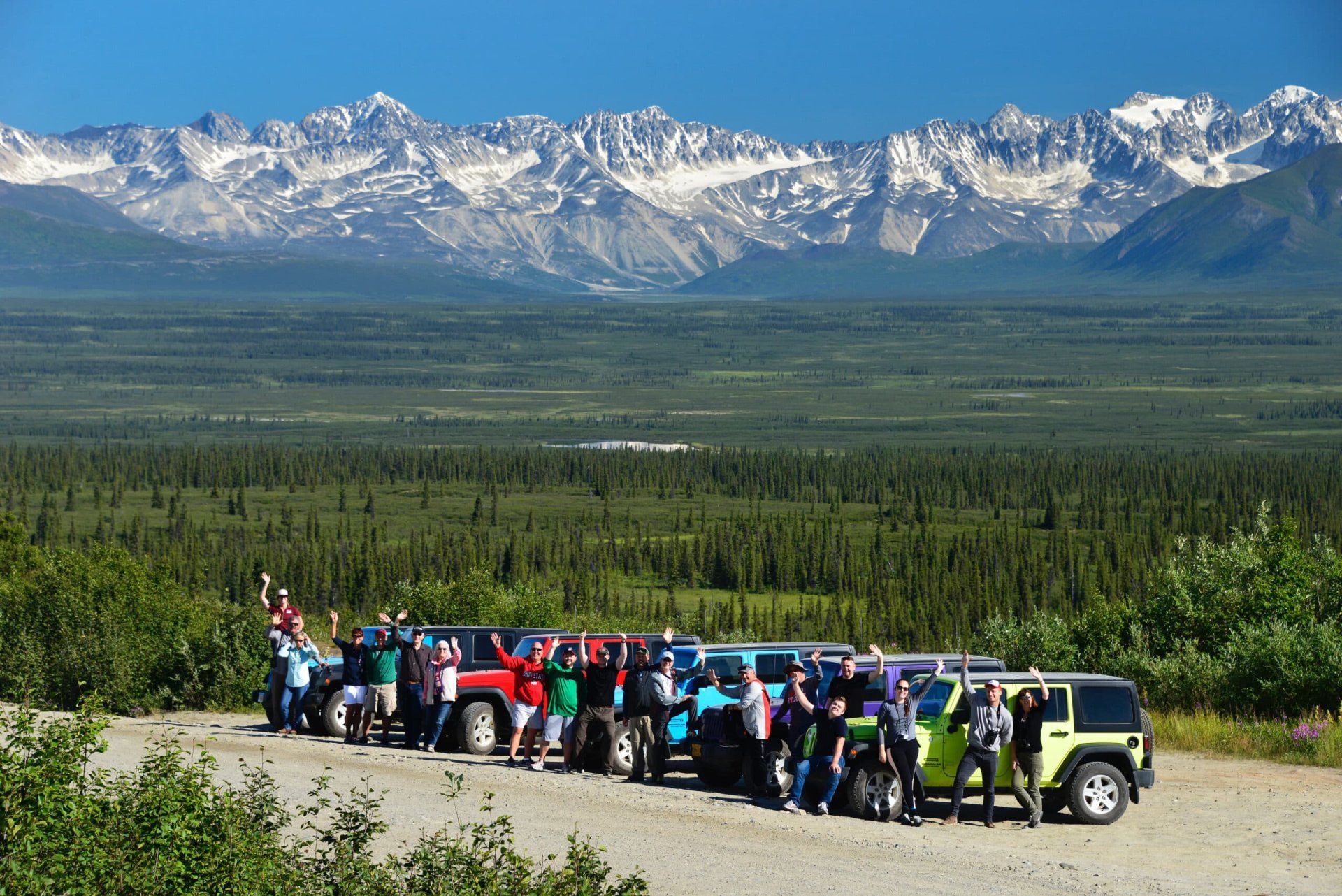 People gathered beside colorful off-road vehicles with snowy mountains and forest in the background