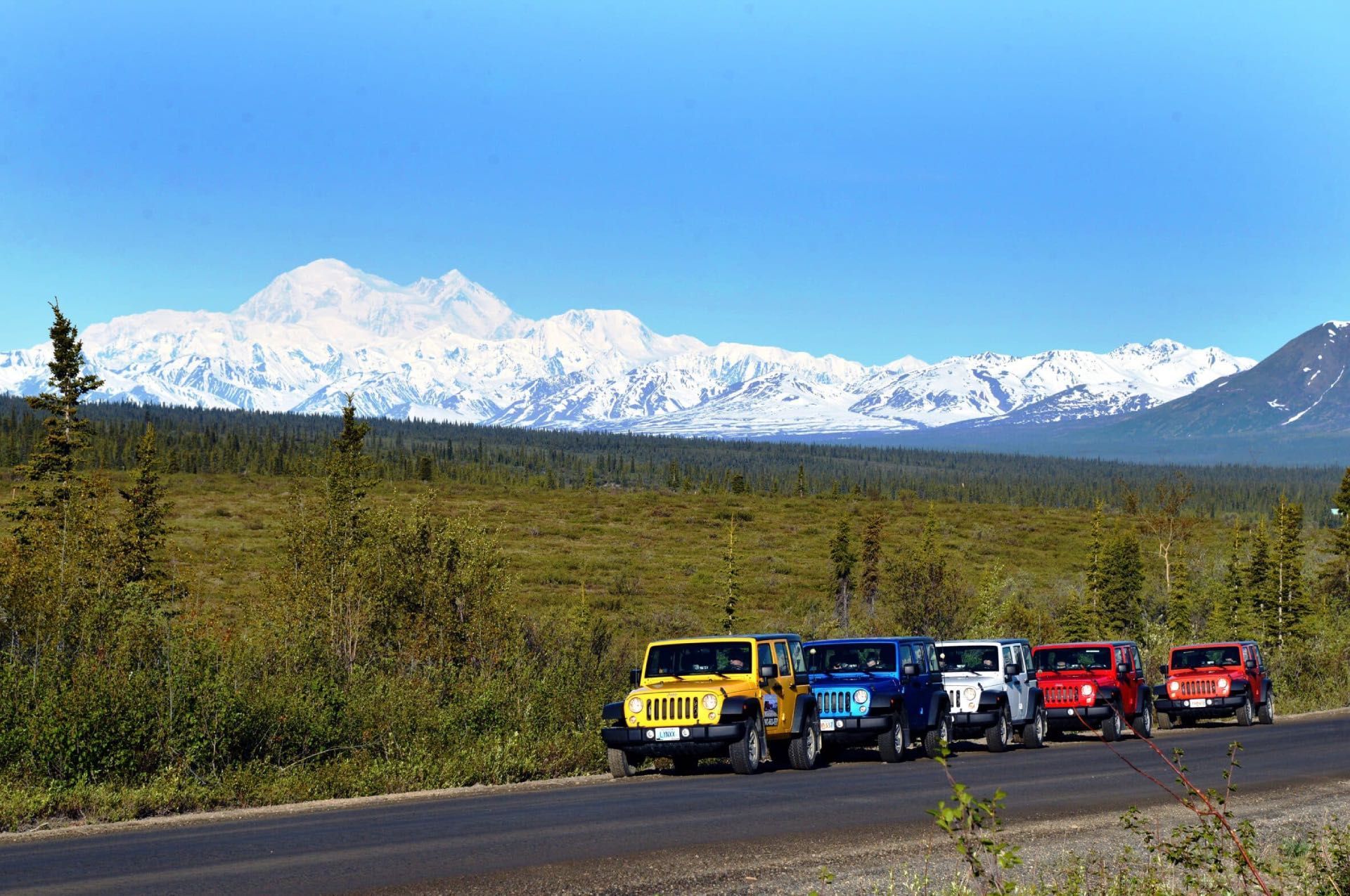Colorful off-road vehicles lined up on a road with snowy mountains and blue sky in the background