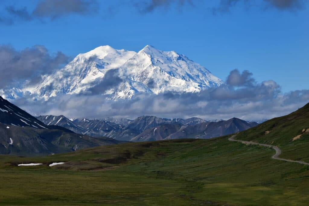 Snow-covered mountain under a blue sky above green rolling hills