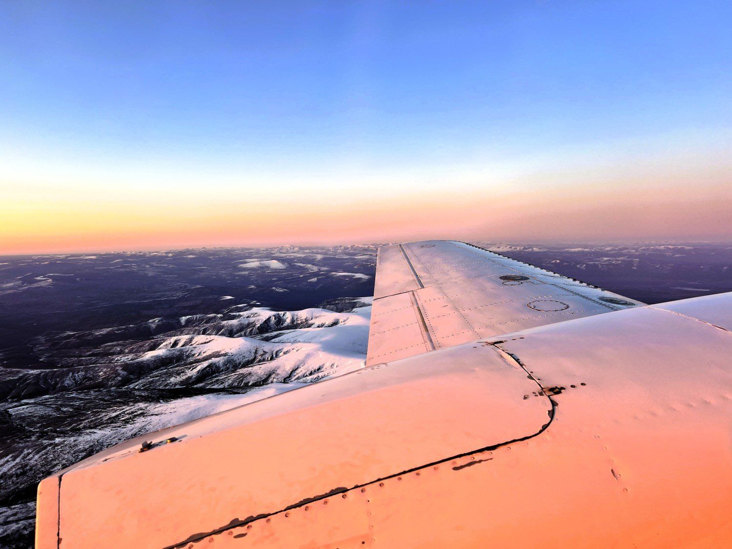 Airplane wing at sunset above clouds, with pink-orange light and a blue sky horizon