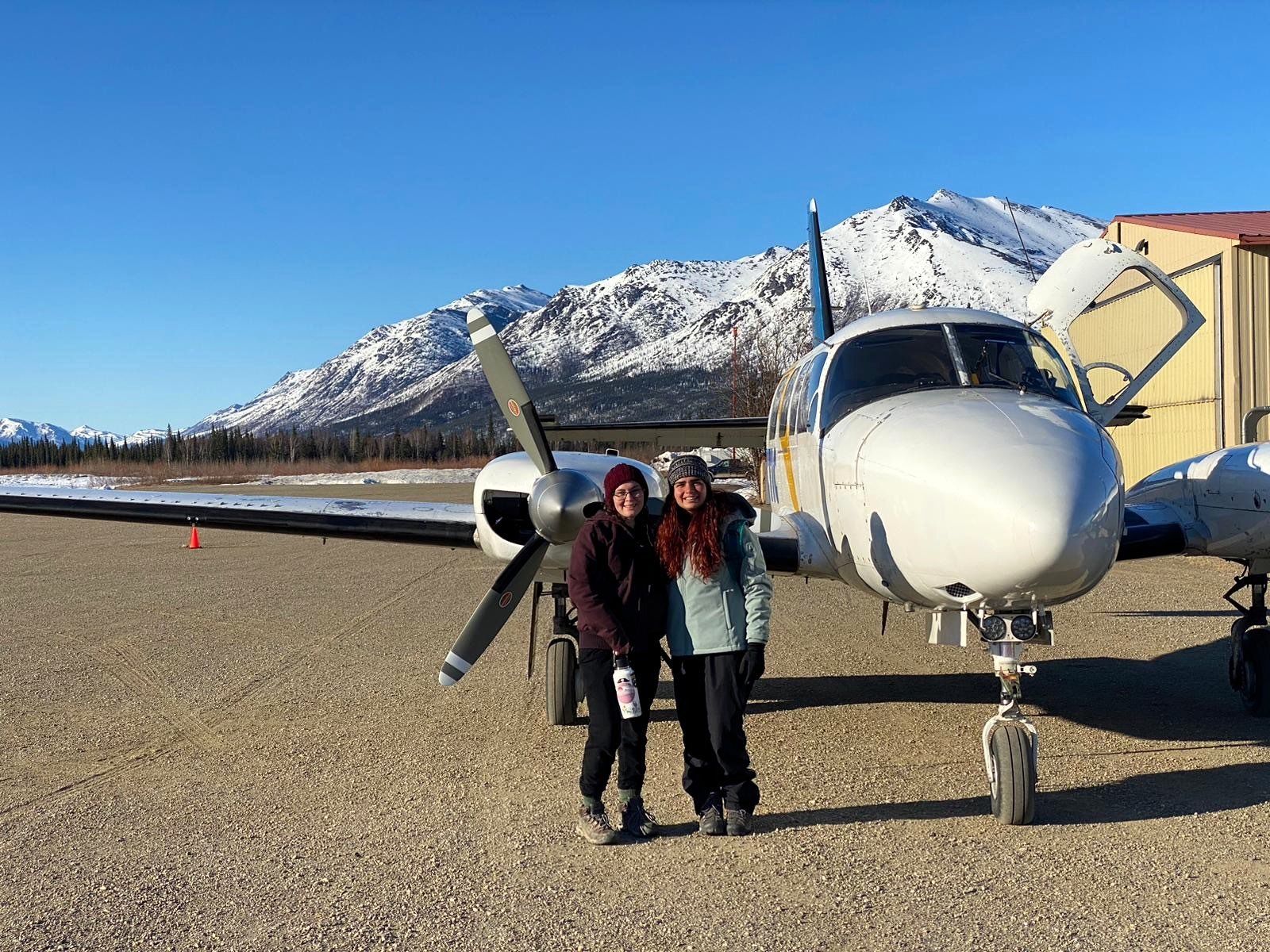 Two people standing beside a small prop plane on a snowy runway with mountains in the background
