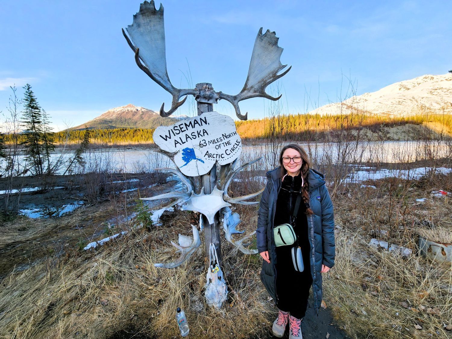 Woman standing beside a moose sculpture in a snowy mountain landscape, holding a sign.