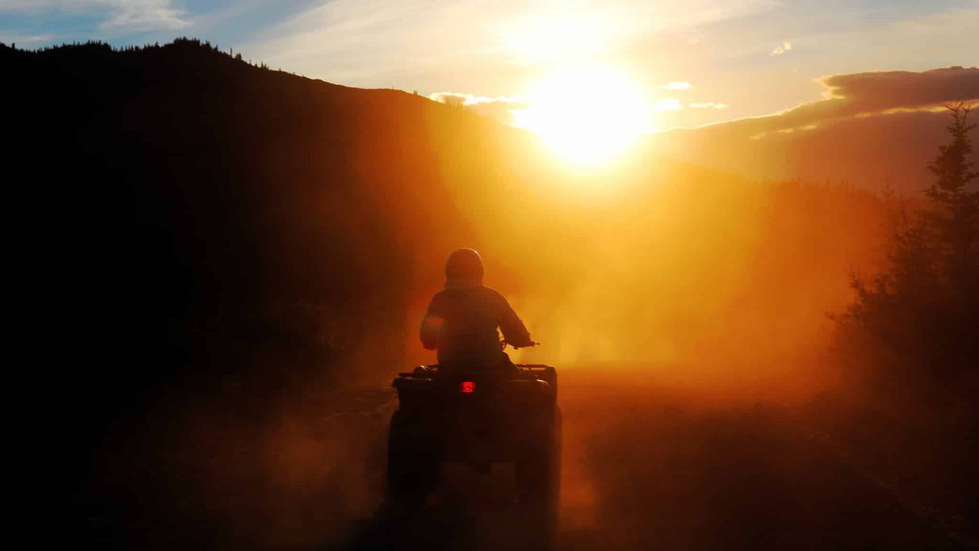 Silhouetted person riding an ATV on a dusty trail at sunset, with bright sun and trees in the background