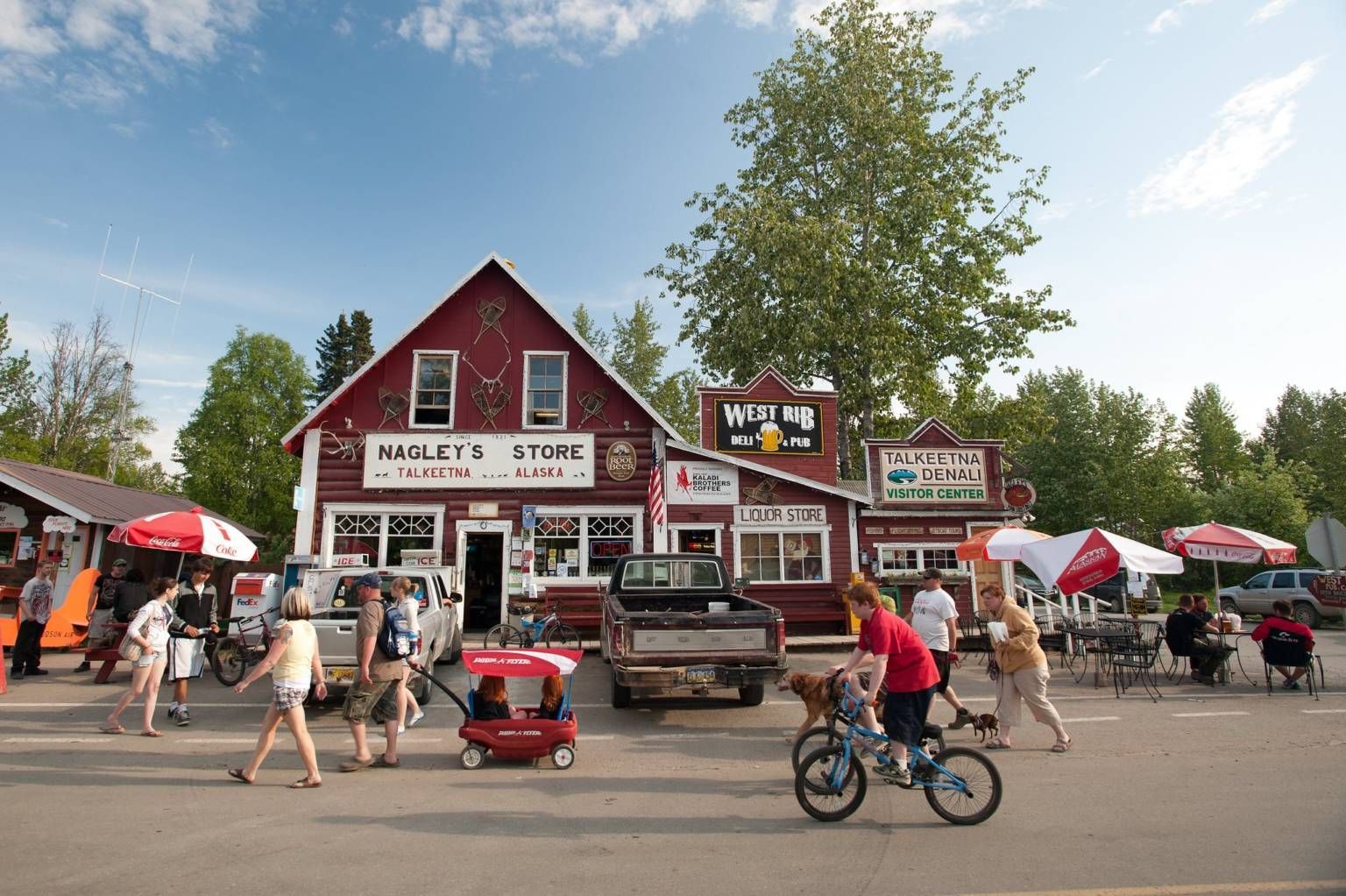 Crowded outdoor market with a red barn-style store, people walking, bikes, and umbrellas on a sunny day
