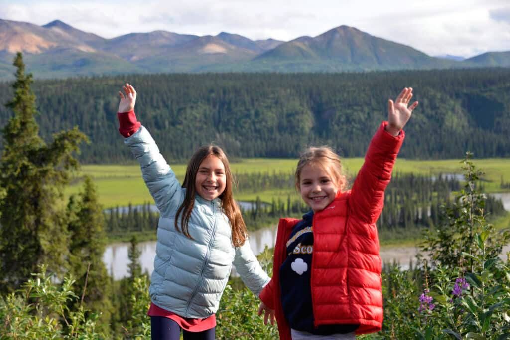 Two children in red and blue jackets waving in a mountain meadow with forest and peaks behind them