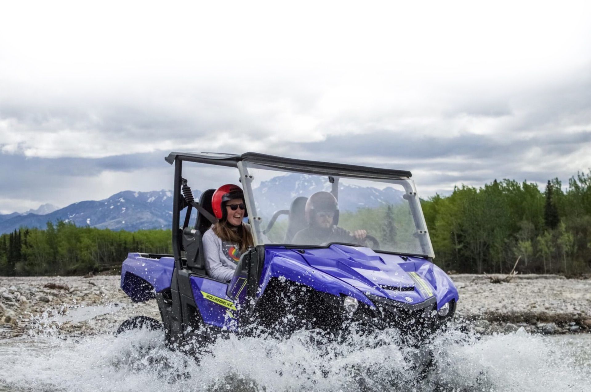 Blue side-by-side ATV splashing through shallow water with two riders in a cloudy mountain landscape