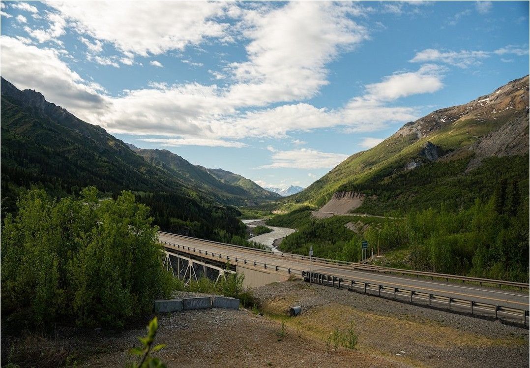 Mountain valley with a winding river, forested slopes, and a road under a cloudy blue sky