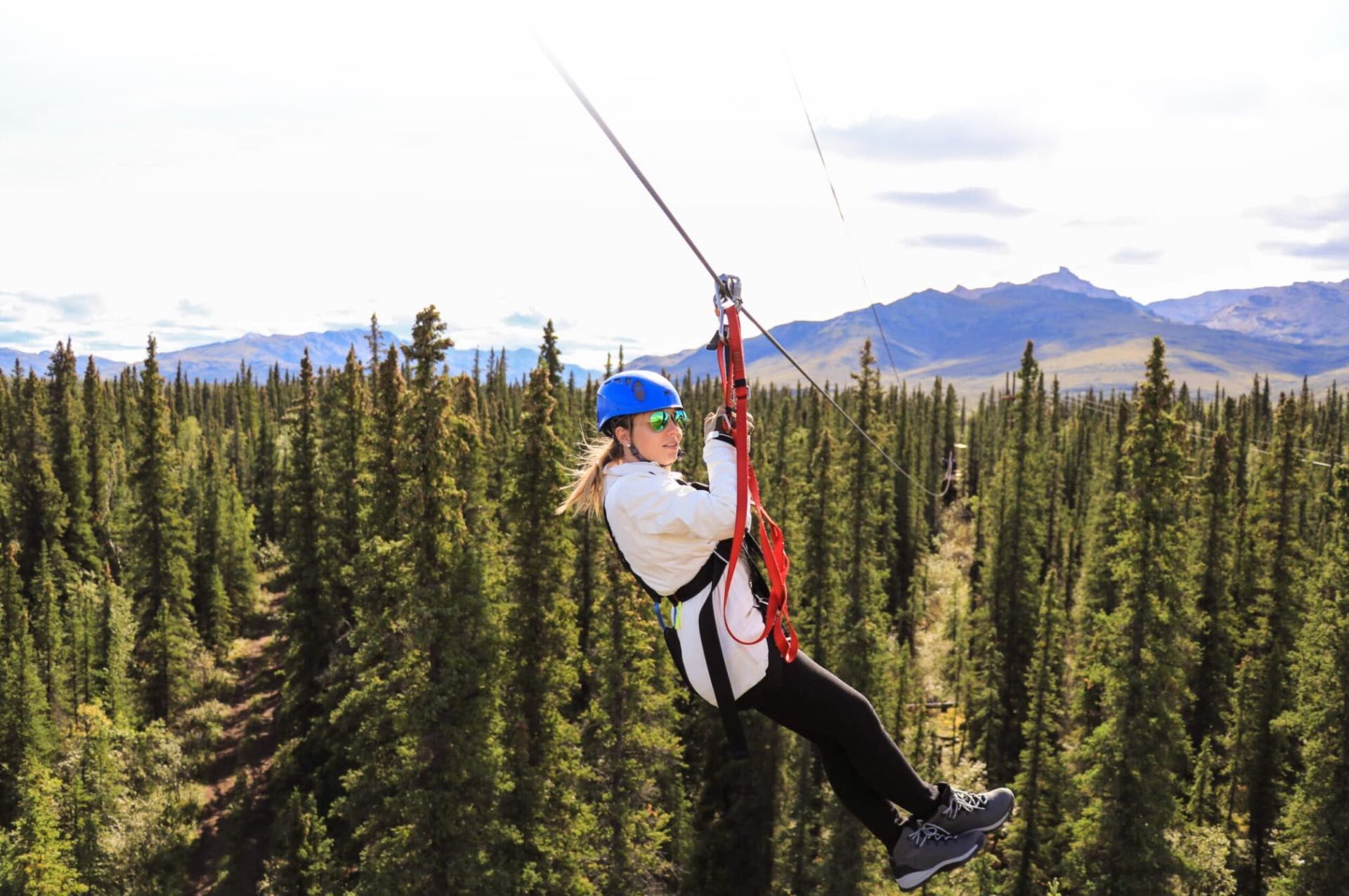 Person zip-lining above a forest, wearing a blue helmet and white jacket with mountains in the background