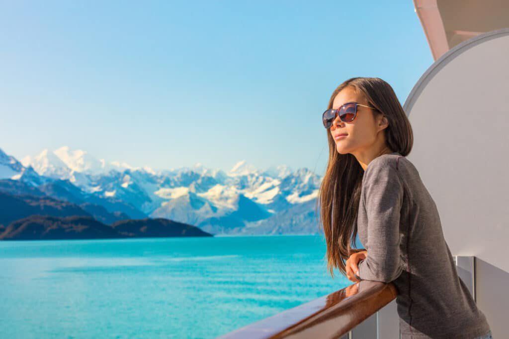 Woman in sunglasses leaning on a boat railing, with turquoise water and snowy mountains in the background.
