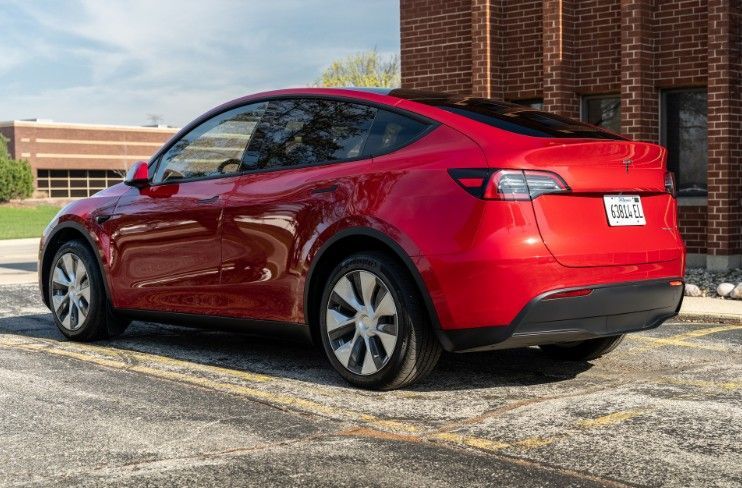 Red Tesla Model Y parked on a street beside a brick building, viewed from the rear side.
