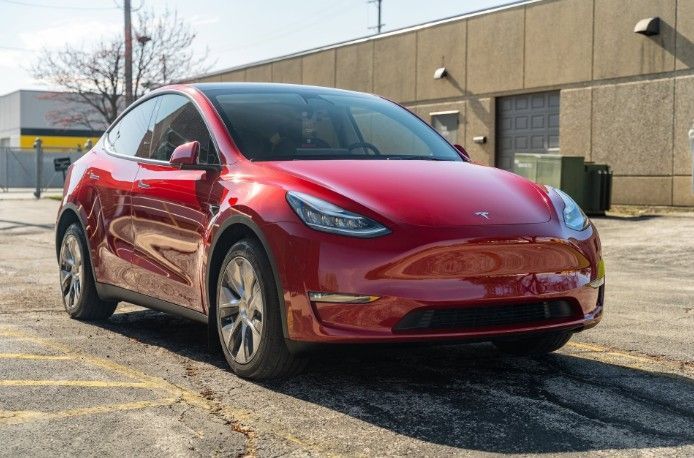 Red Tesla Model Y parked on a street beside a beige building, viewed from the front-left.