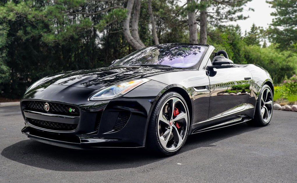 Black Jaguar F-Type convertible parked outside, shiny paint, red brake calipers, bright sunlight, trees in the background.