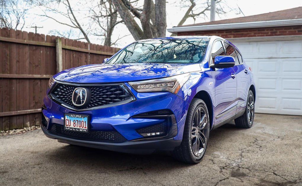 Blue Acura RDX parked in front of a garage, in a driveway. Dark gray wheels, brown fence, and overcast sky.