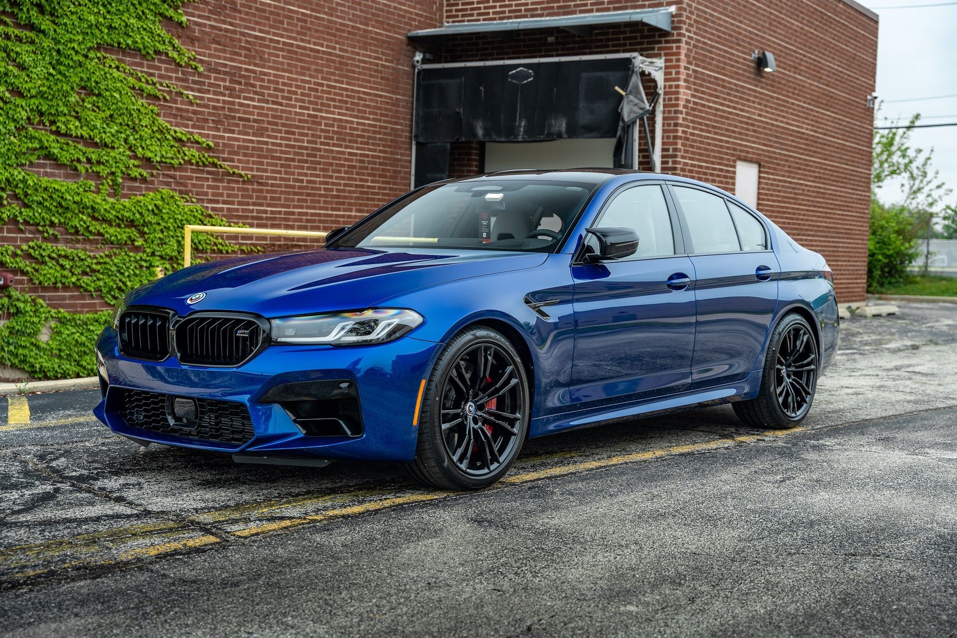 Blue BMW M5 sedan parked on asphalt, in front of a brick building with a garage door open.