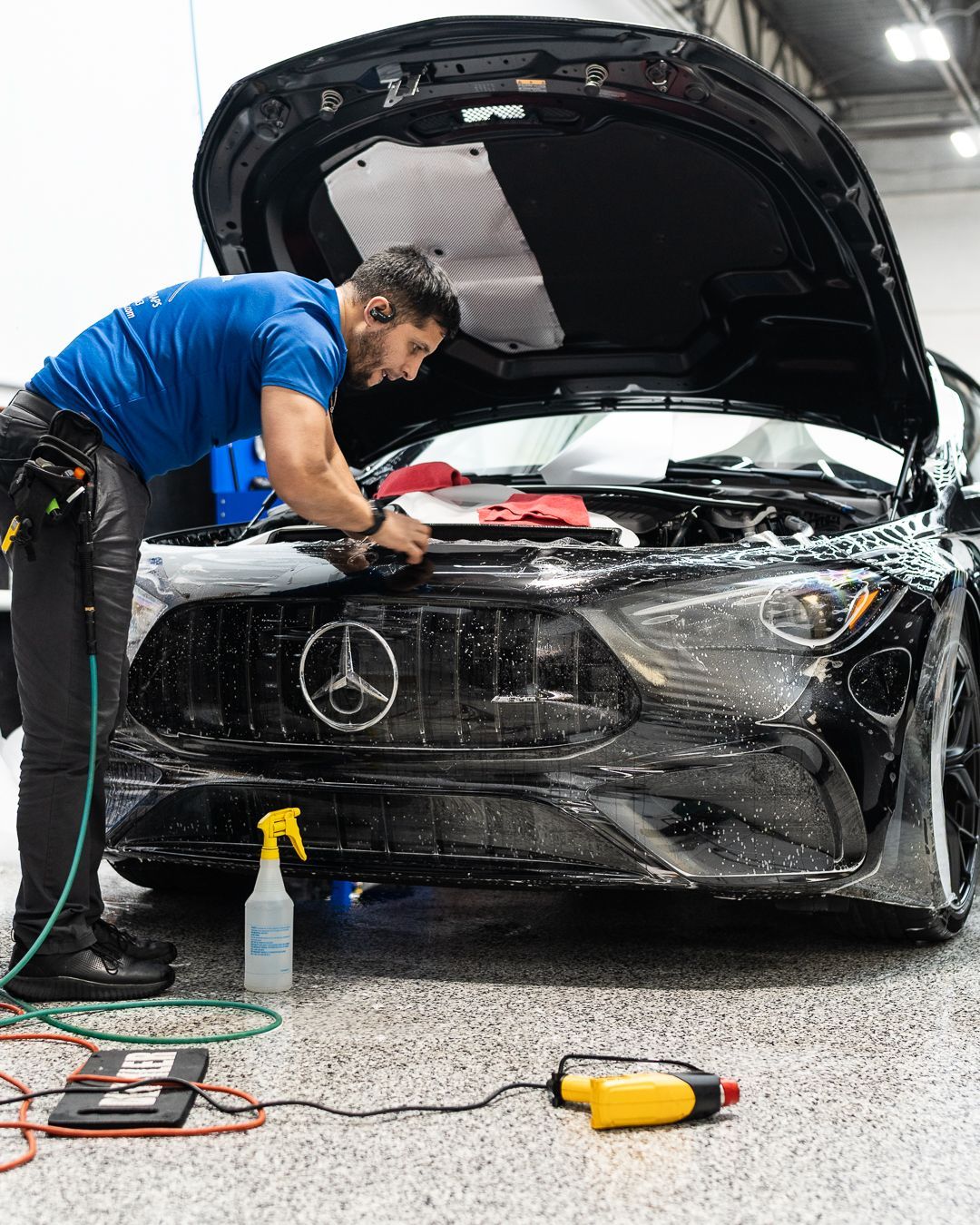 Mechanic polishing a black Mercedes in a garage, hood open, with tools and spray bottle nearby