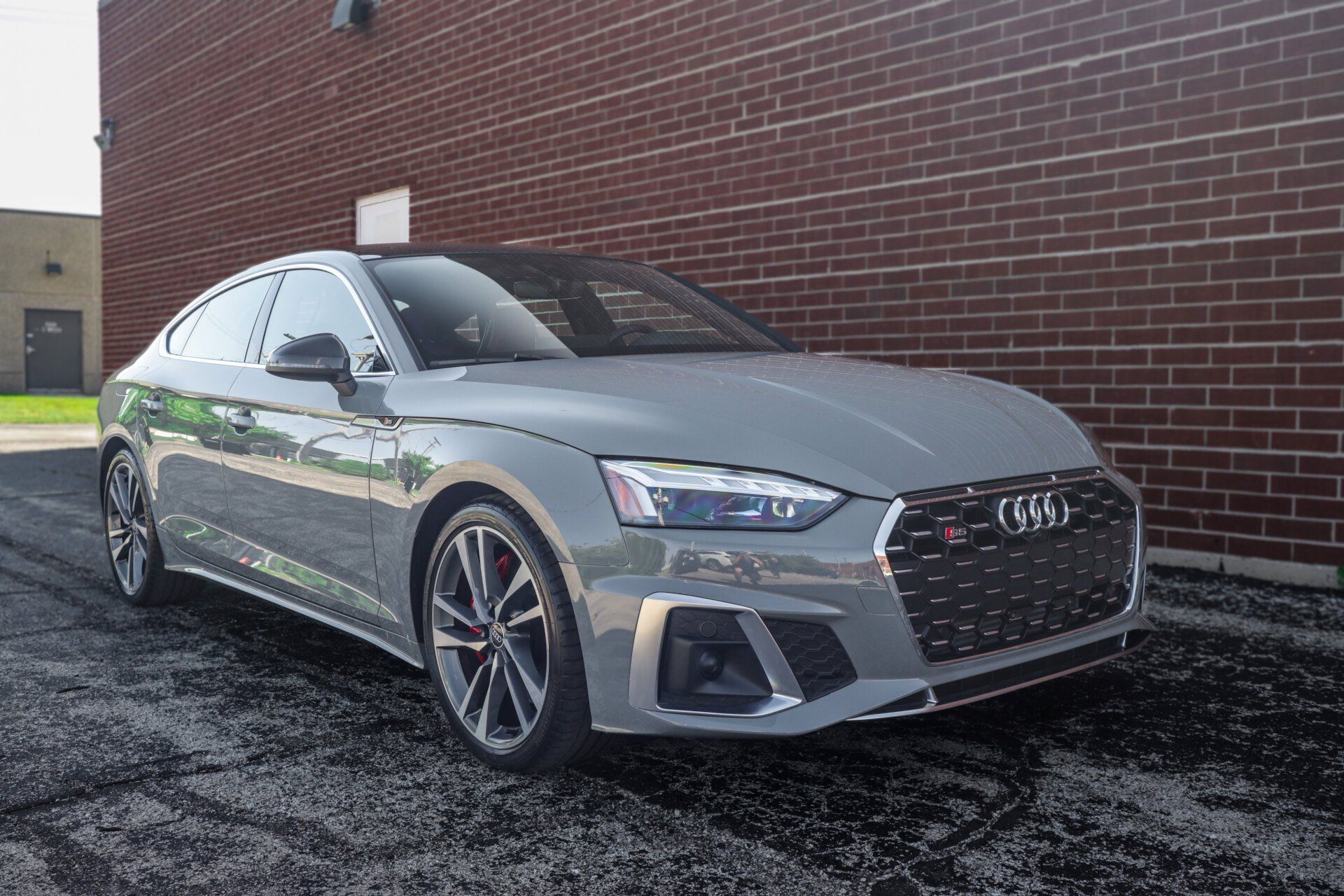 Gray Audi S5 coupe parked on asphalt in front of a red brick building.