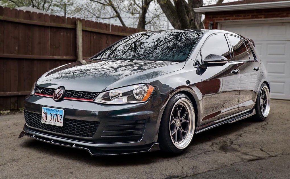 Gray Volkswagen GTI hatchback with custom wheels and front lip, parked in front of a garage.