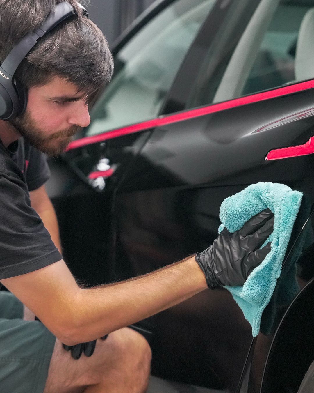 Man in headphones, black gloves, wiping black car panel with a blue cloth.