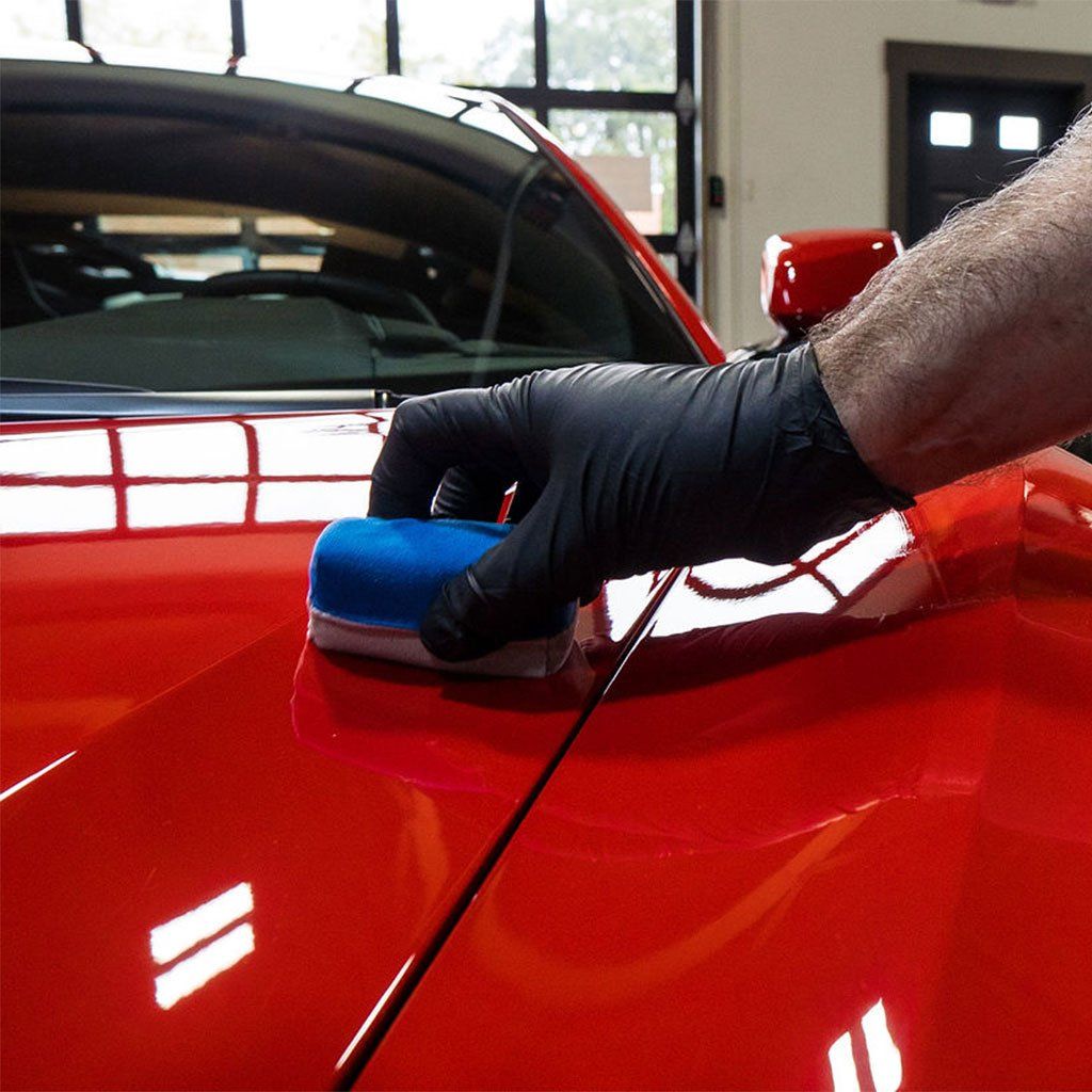 Gloved hand polishing a red car hood with a blue applicator in a garage