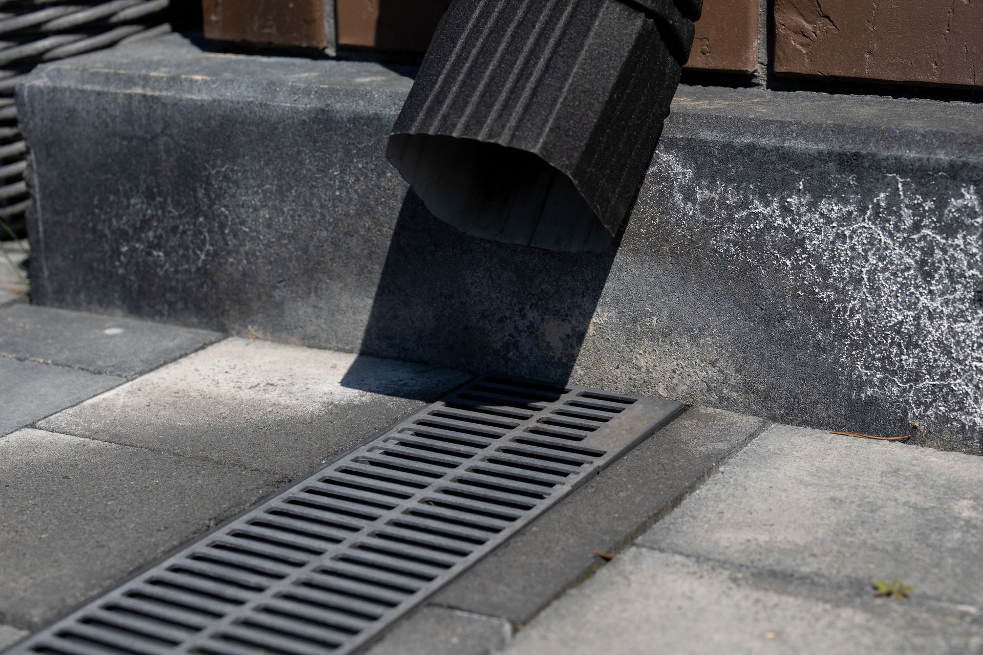 Black metal downspout discharging rainwater onto a paved walkway beside a drainage grate.