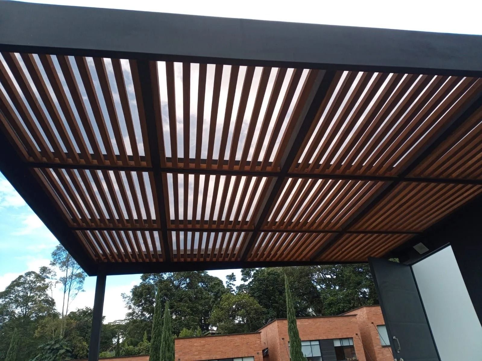 Pérgola con techo de listones de madera sobre un patio exterior con árboles y un edificio de ladrillo al fondo.
