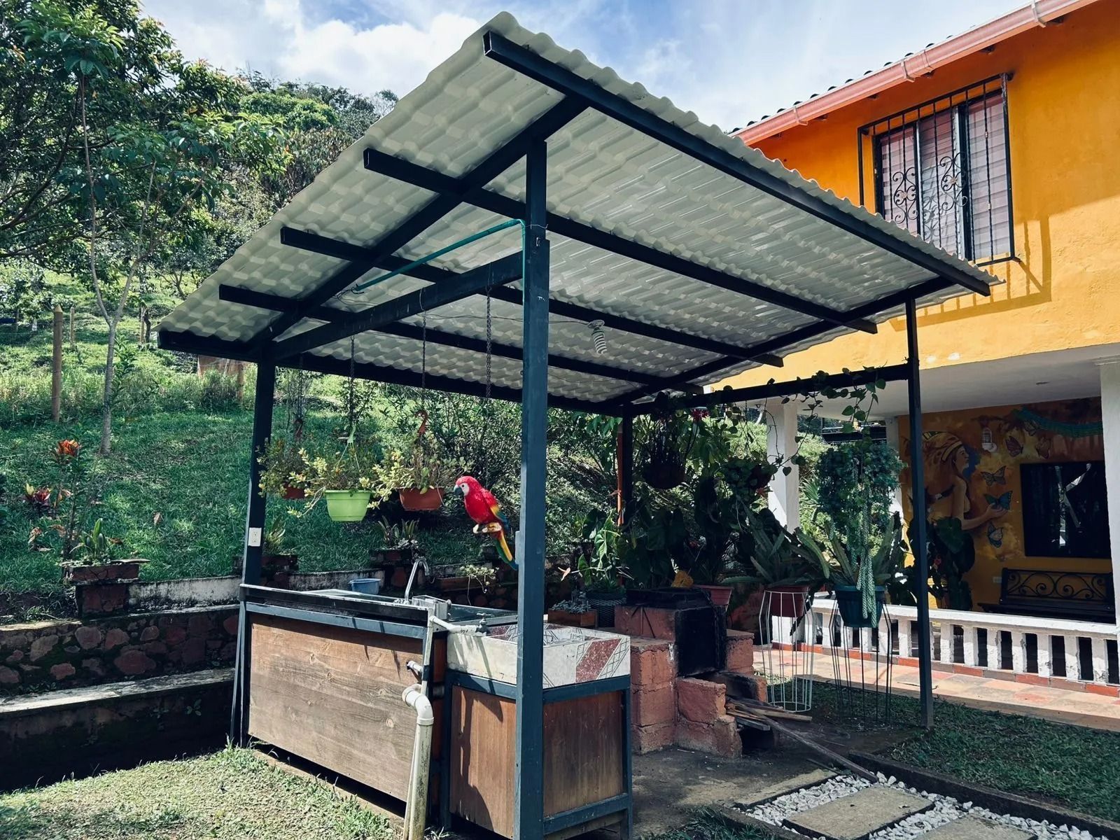Patio exterior cubierto con techo de metal, plantas en macetas y una casa amarilla junto a una ladera verde.