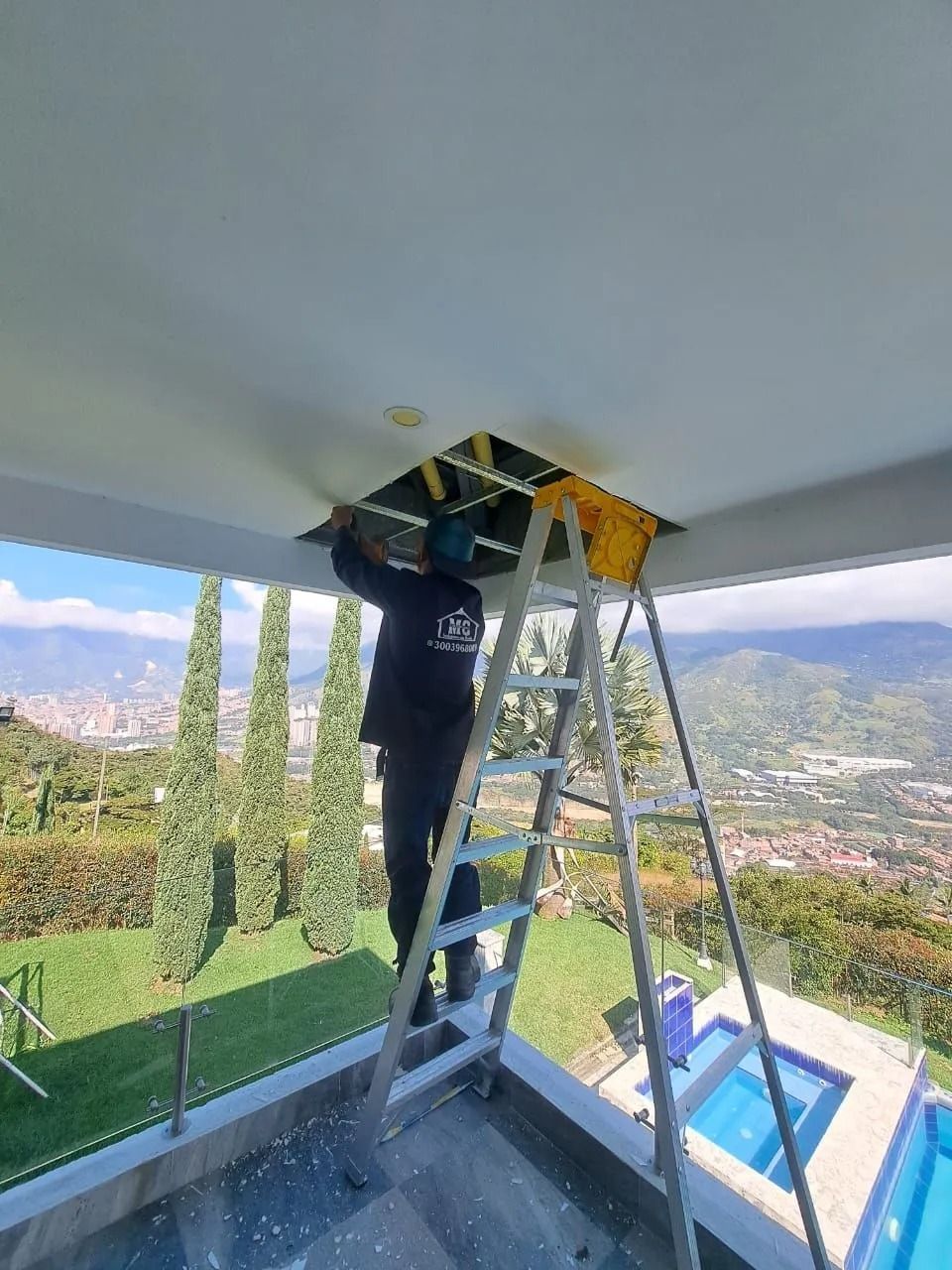 Una persona subida a una escalera repara un panel del techo de un balcón con vistas a un valle de montaña.