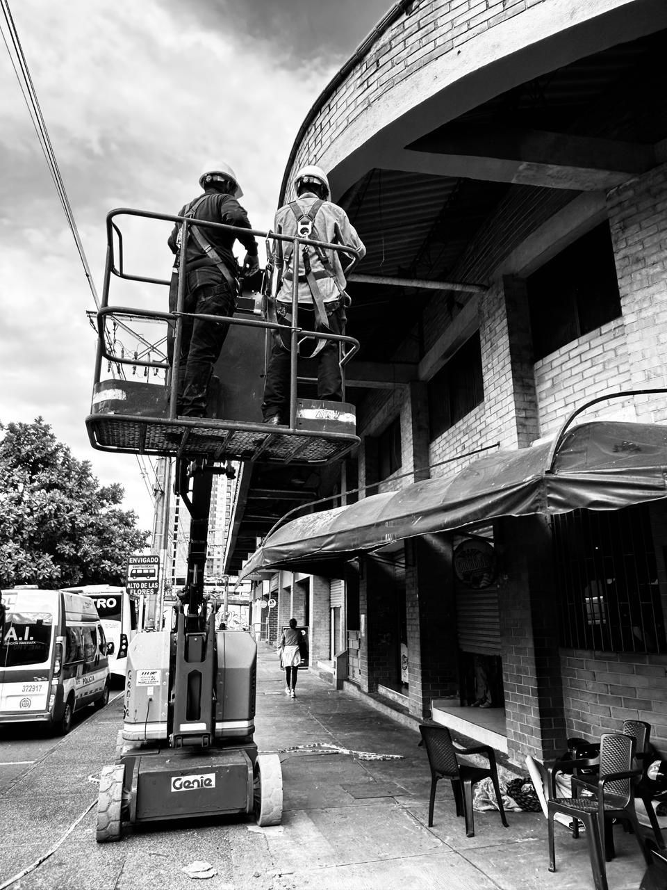 Dos trabajadores en un ascensor repintando un edificio de ladrillo con toldos arqueados.