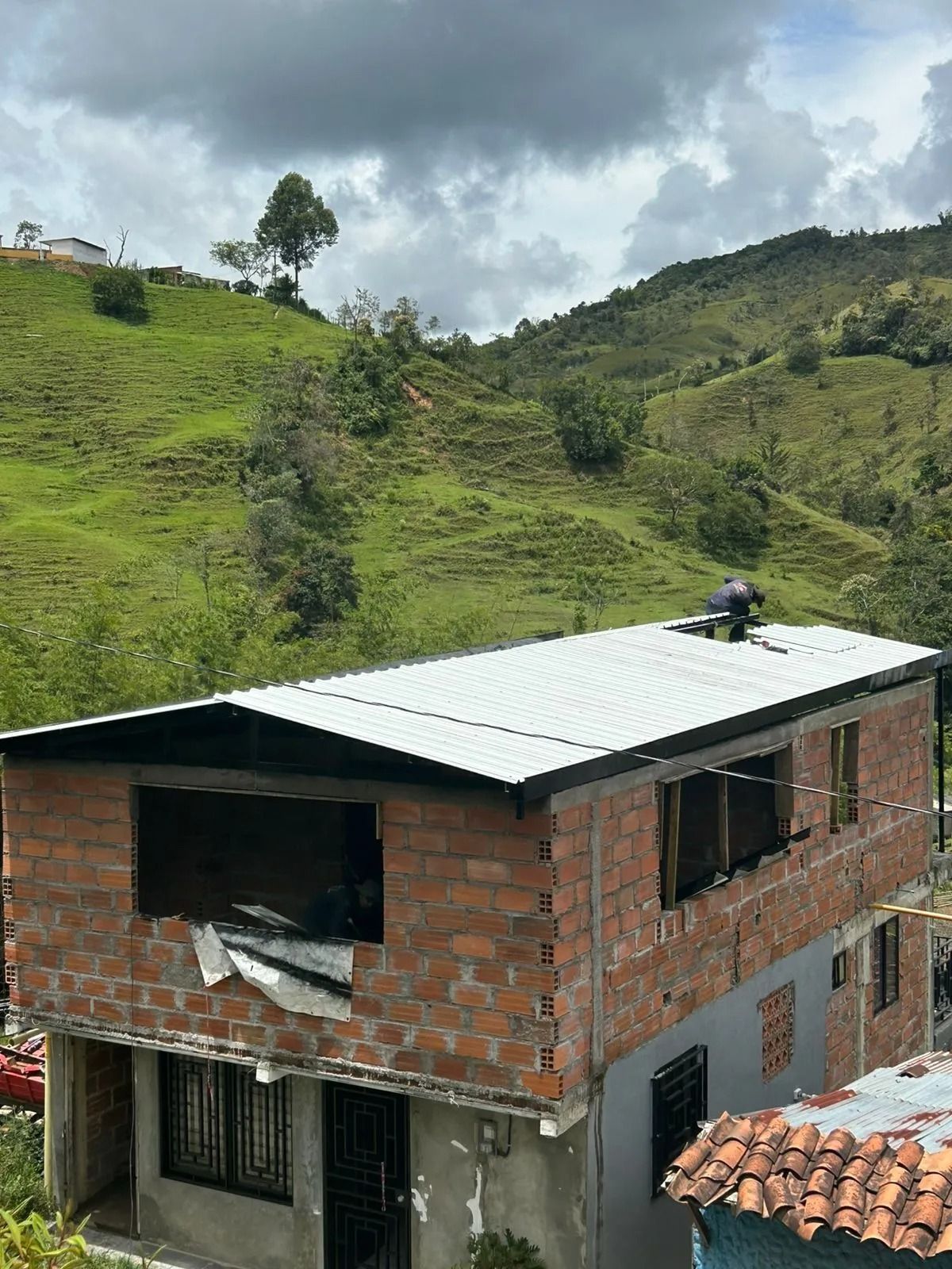 Casa de ladrillo sin terminar con techo de metal en una ladera verde bajo un cielo nublado.
